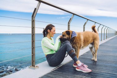 30 'lu yaşlardaki mutlu beyaz kadın sahilde en iyi arkadaşla vakit geçiriyor. Morgel köpekli, spor giyinen neşeli bayan antrenör sevimli evcil hayvanlarla iletişim kurarken gülüyor.