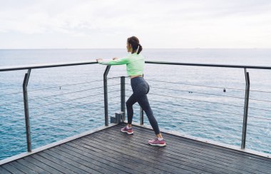 Beyaz kadın sporcu vücut çalıştırıyor kasların gücünü arttırıyor, kıyı şeridi koşusuna hazırlanan kararlı sporcu sabah esneklik egzersizleri yapıyor ve fitness hedeflerini koruyor.