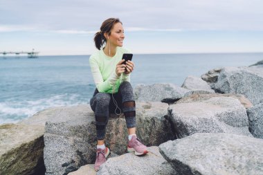 Deniz taşlarında dinlenen spor eşofmanlı neşeli beyaz koşucu motivasyon müzik podcastının keyfini çıkarıyor, akıllı telefon teknolojisine indirilen olumlu ses şarkılarını dinleyen mutlu formda kız.