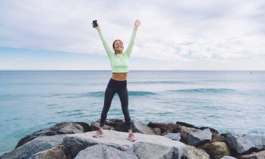Cep telefonuyla spor zaferi ve koşu golüyle tatmin olan mutlu Kafkasyalı fitness kızı, kulaklıklı kaygısız bir kadın sahil taşlarında el kaldırırken müzik dinliyor.