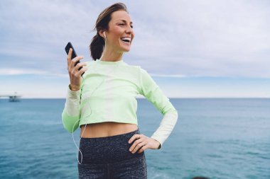 Kulaklık takmış, deniz manzarasının tadını çıkaran ve deniz kıyısında dinlenirken cep telefonuyla müzik dinleyen neşeli, sıska bir genç bayan.