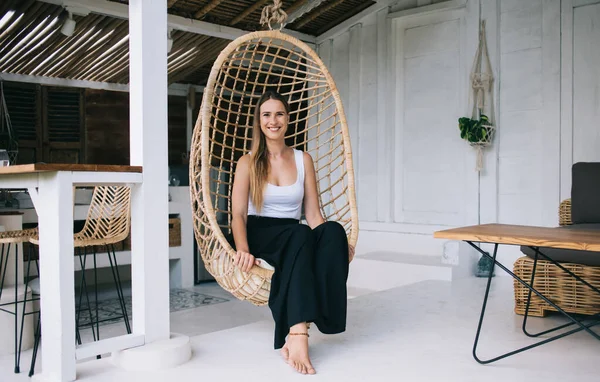 Full body of cheerful young woman in casual clothes smiling and relaxing on wicker hanging egg chair in cozy house