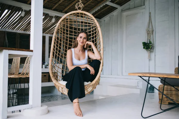 Beautiful friendly lady in causal clothes enjoying peace and quiet of summer terrace while relaxing in wicker hanging egg chair