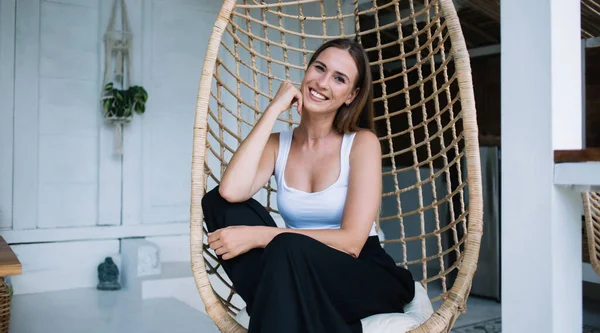 Cheerful attractive lady in casual white summer top and black wide trousers sitting in wicker hanging egg chair and looking at camera with broad smile