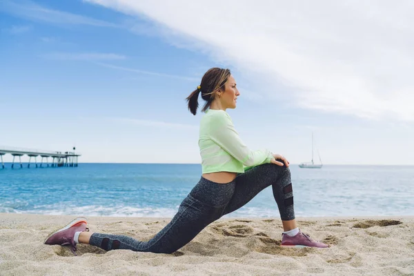穿着舒适运动服的柔韧女健美运动员蹲着做伸展运动以热身腿肌肉的侧视图 白种人适合的女孩在奥西亚海岸享受晨练 — 图库照片