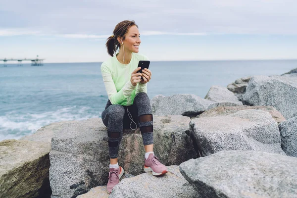Cheerful Caucasian jogger in sportive tracksuit resting at seashore stones enjoying motivation music podcast, happy fit girl listening positive audio songs downloaded to smartphone technology