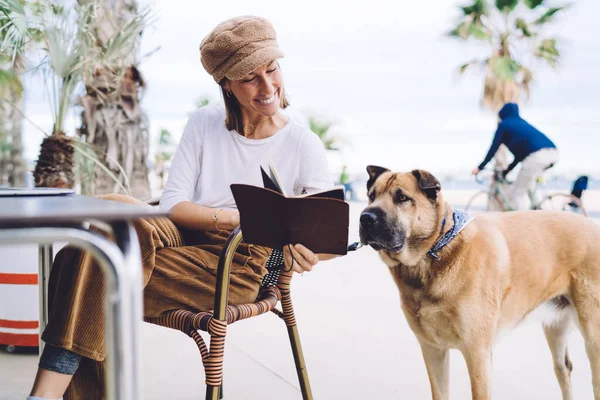 Happy pretty woman journalist satisfied with creative article sitting on cafe terrace during free time, smiling caucasian female in trendy wear having fun with lovely puppy dog showing notebook to pet