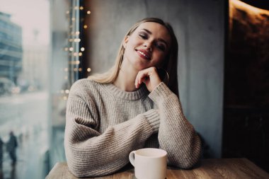 Kafkas modeli, eğlence zamanı kameraya gülümsüyor aromalı kafein içeceği içiyor, neşeli kız kafeteryada çay fincanı ile masada oturarak ısınmanın keyfini çıkarıyor.