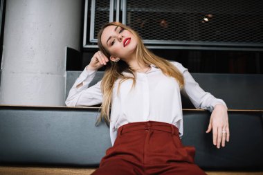 Modern karanlık bir yerde rahat deri koltukta oturan ve kameraya bakan modaya uygun giyinmiş ciddi ve şık genç bir kadının alçak açısı.