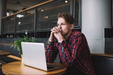 Giysileri gündelik giyinmiş yakışıklı erkek öğrenci ellerini birbirine kenetleyerek kafede otururken dikkatle başka tarafa bakıyor.