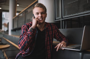 Modern, rahat kafeteryalarda çalışırken kameraya bakıp, akıllı telefondan konuşan rahat ve rahat genç bir erkek.