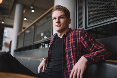 Kafedeki deri koltukta oturan, güpegündüz kafasını çeviren, kırmızı kareli gömlekli, kendine güvenen sarı saçlı adam.