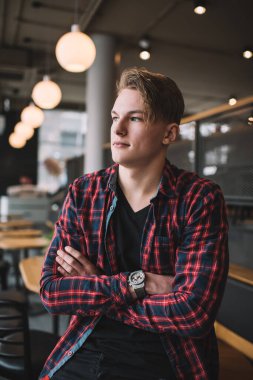 Gerçekten yakışıklı, sıradan giyinmiş, yaratıcı saç stili olan, kollarını kavuşturmuş, modern bir kafede durup düşünceli bir şekilde başka tarafa bakan bir adam.