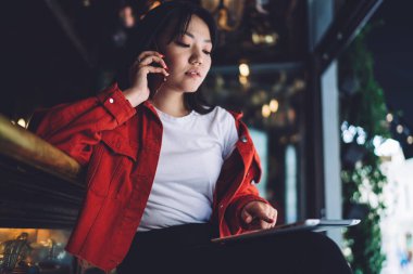 Barda otururken günlük giysiler giyen, cep telefonuyla konuşan ve tablet okuyan genç etnik kadın bağımsızlardan.