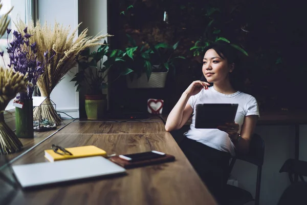 白いTシャツを着たアジア系の女性のフリーランサーが スマホを持った木製の机に座って遠隔作業中に装置を使いながら目を離している — ストック写真