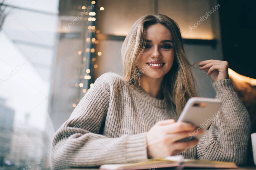 Portrait of cheerful hipster girl with perfect veneers smiling at camera while using smartphone device, funny female student with education textbook posing while learning and blogging indoors