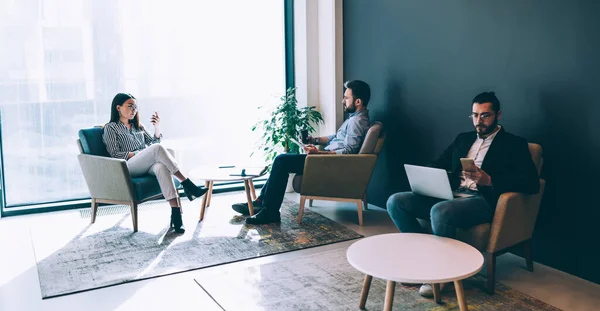 Businessman in glasses with laptop on knees using smartphone and sitting next to dark haired man who looking at big window and focused woman