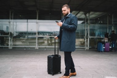 Kafkasyalı yolcu, havaalanının dış cephesinde modern cep telefonu cihazıyla elektronik bileti kontrol ediyor. Resmi olarak giyinmiş, bagaj taksisi sipariş eden bir iş adamı.