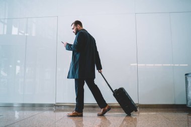 Havalimanında bavulla yürürken cep telefonundan e-postaları kontrol eden şık bir işadamı, iş seyahatinden dönüş biletleri için cep telefonu teknolojisi kullanan resmi giyinmiş bir erkek işveren.