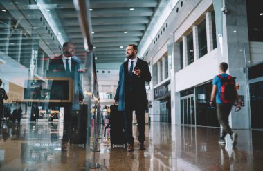 Modern havaalanı terminalinde gülümseyen ve elinde telefon ve bavulla yürüyen resmi kıyafetli mutlu genç işadamlarıyla dolu.