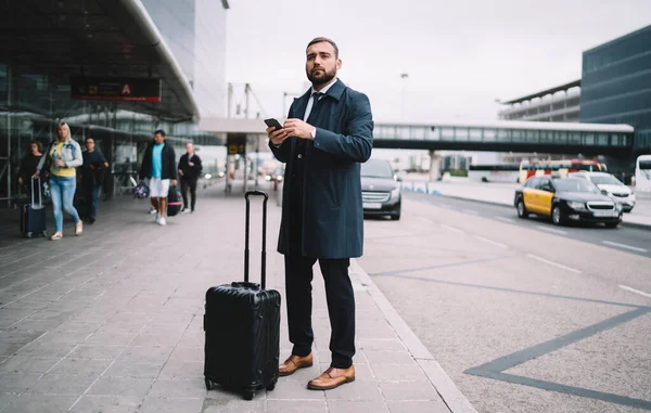 旅行中の出発を考慮して空港近くに立つ正式な衣服の男性従業員 携帯電話技術を保持し 端末を探しているスーツケースの荷物を持つ白人起業家 — ストック写真