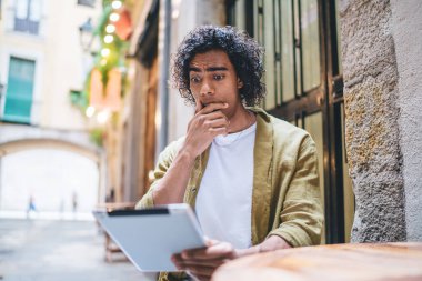 Düşük açı, sokak kafesinde vakit geçirirken tablette ilginç hikâyeler okuyan sıradan, genç Afrikalı Amerikalı bir erkek.