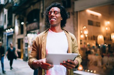 Sıradan giyinen neşeli yakışıklı hippi, dijital tabletle şehir caddesinde duruyor. Blogda seyahat hakkında bilgi paylaşımı yapıyor. Gülümseyen erkek, dokunmatik dokunmatik uygulama ve iletişim kurmayı seviyor.