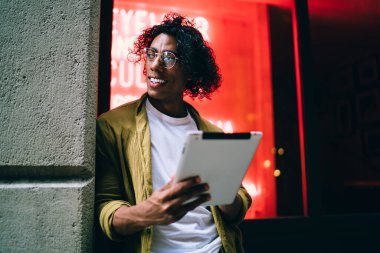 Gülümseyen kıvırcık kıpır gözlüklü, neon ışıklı sokakta dikilip dijital tablet paylaşımı yapan yakışıklı adam, bloglarda touchpad kullanan ve medyayı ve multimedya içeriğini paylaşan adam.