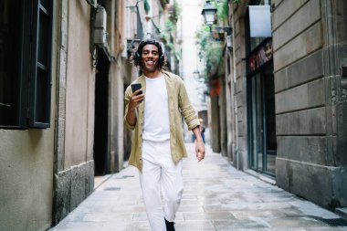 Zeki telefonlu, modaya uygun kıyafetli, kendine güvenen, kulaklıkla müzik dinleyen, güler yüzlü, dar bir sokakta yürüyen genç bir adam.