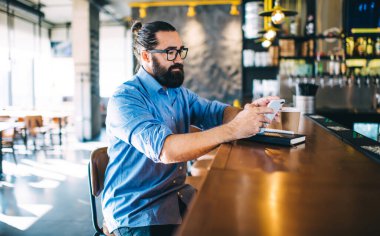Odaklanmış orta yaşlı, uzun siyah saçlı, rahat giyimli ve gözlüklü, akıllı telefon kullanan, gündüzleri kafenin tezgahında oturan hippi bir erkeğin yan görüntüsü.