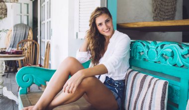Genç, esmer, kıvırcık saçlı, dişlek tebessümlü, turkuaz bankta oturan, rahat verandada çizgili yastıklı bir kadın.