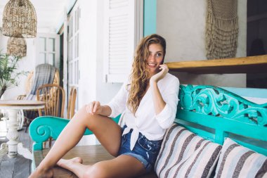 Gülümseyen, güzel saçlı, neşeli bir kadın akıllı telefonda konuşuyor rahat bir poz veriyor açık havada geniş bir bankta oturuyor.