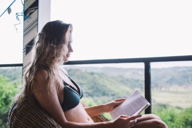 Siyah bikinili, düşünceli, düşünceli bir kadının yaz mevsiminde terasta hasır koltukta dinlenirken ilginç bir kitap okuması.