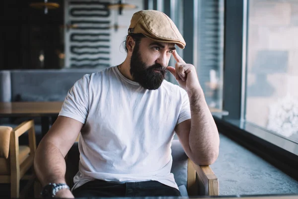 Thoughtful Bearded Man Trendy Cap Casual Clothes Sitting Wooden Chair — Stock Photo, Image