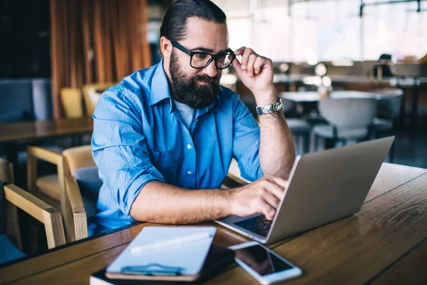 Adult beared brutal man with glasses in wrinkled clothes using laptop while sitting at creative workplace and working on project