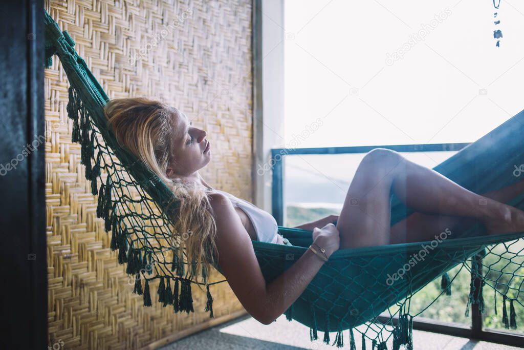 Side view of relaxed tanned woman in swimsuit resting resting and sleeping in hammock in open terrace with garden access on bright sunny day