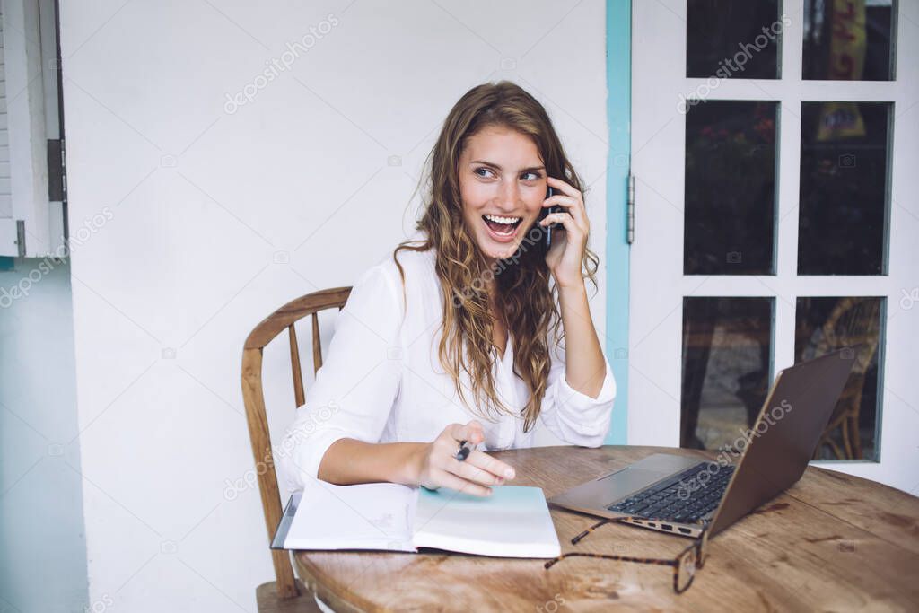 Agreeably surprised curly red haired lady in casual clothes gossiping with friend on mobile phone with big opened smile and looking away while working with laptop at terrace 