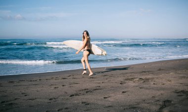 Güneşli yaz gününde sörf tahtasıyla dalgalanan kumlu sahilde mayo giymiş neşeli sarışın bir kadın.