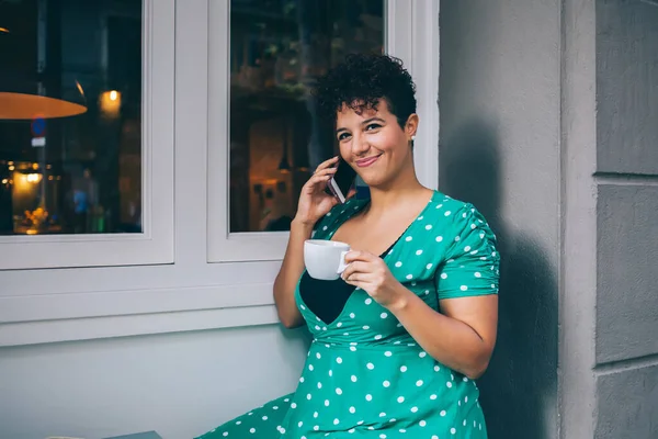 Portrait of cheerful woman in trendy dress enjoying mobile connection for making online international phoning vie cell technology, happy Hispanic female 20s with caffeine beverage calling in roaming