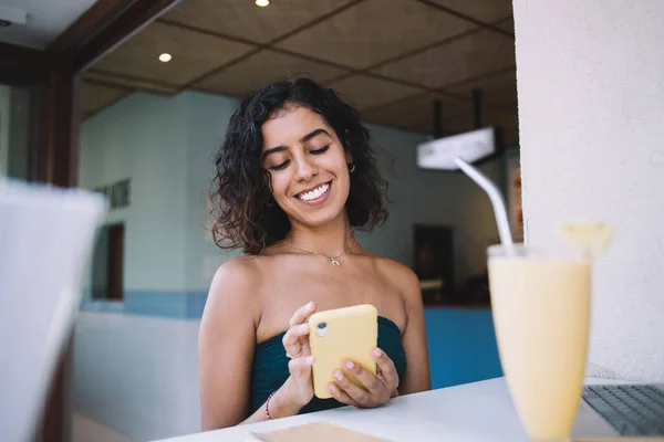 Cheerful Middle Eastern woman using cellphone gadget for writing feedback during bistro visiting, happy female customer with milkshake on frontage writing sms message during smartphone chatting