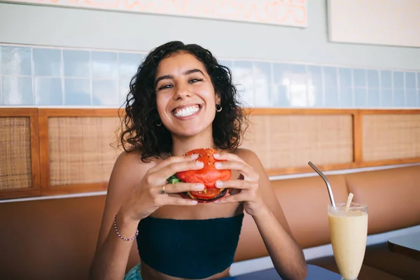 Portrait of joyful female customer with cheeseburger smiling at camera during cheat meal day in bistro cafeteria, funny Middle Eastern hipster girl with tomato bread rolls enjoying American dinner
