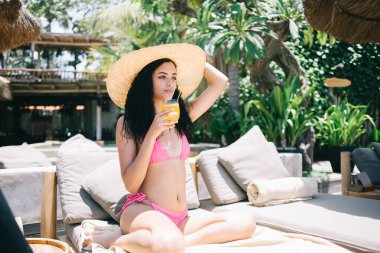 Pembe bikinili, hasır şapkalı, meyve kokteyli içen ve tropik tatil beldesinde güneşlenirken yüzünü çeviren genç etnik kadın.
