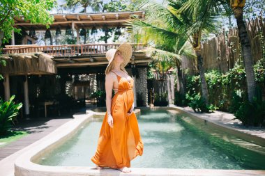 Güzel, sarışın, şapkalı hamile bir kadın tropikal villa tatil köyünde boş vakitlerin tadını çıkarıyor. Lüks elbiseli genç bir kadın hafta sonları lüks otellerde havuz kenarında yürüyor.