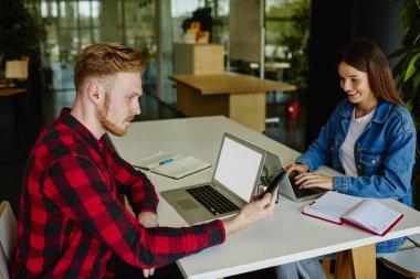 Kafkasyalı erkek programcı, masa başında mutlu bayan meslektaşıyla birlikte internet reklamı için fotokopi alanı olan sahte dizüstü bilgisayarı kullanarak mobil cihazdan gelen e-postayı kontrol ediyor.