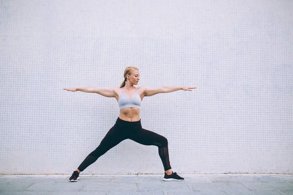 Energetic slim woman in tracksuit doing Warrior one during yoga exercises in morning, female gymnastic enjoying virabhadrasana during time for physical recreation at publicity area with copy space