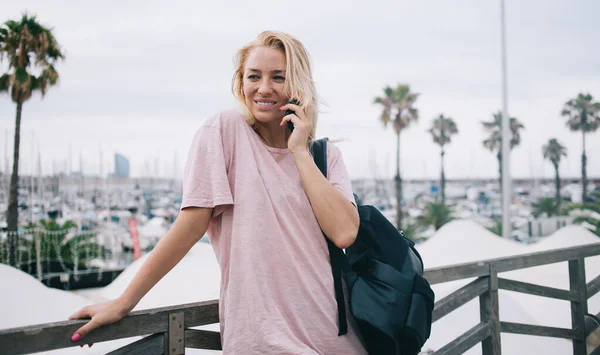 Cheerful female tourist with backpack connecting to roaming internet for calling to best friend and discuss solo travel vacations, happy girl in casual wear enjoying international mobile talking