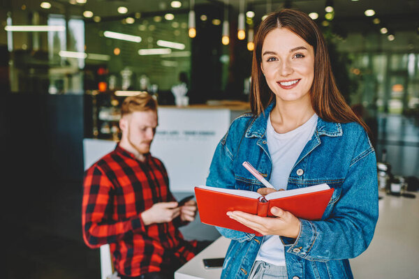 Половинный портрет жизнерадостного кавказского студента, делающего журналистский проект с использованием ручки и учебника, успешной женщины с учебником знаний в руках, улыбающейся в камеру