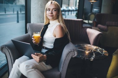 Şık giyinen genç bir kadın dizüstü bilgisayar kullanıyor ve gündüzleri modern kafede otururken taze meyve suyu içiyor.