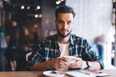 Neşeli, sakallı, damalı tişörtlü, rahatlayan, kahve içen ve gülümseyen bir şekilde kameraya bakan serbest bir erkek. Çatı katı kafeteryasında iş molasında tahta masada gazeteler ve akıllı telefonlarla otururken.