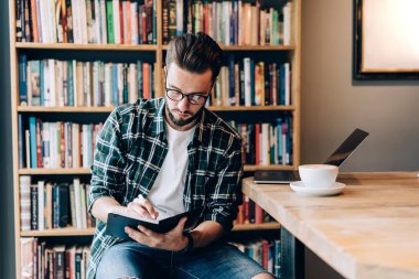 Kafe 'de dizüstü bilgisayarda yazı yazan sakallı genç adam. Duvardan duvara büyük bir kitaplık ve masanın üzerinde bir fincan kahveyle.